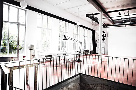 Ein heller, moderner Raum mit großen Fenstern, die viel Licht hereinlassen. Der Raum hat einen roten Boden und verfügt über eine industrielle Gestaltung mit sichtbaren Deckenbalken. Es gibt eine lange Tischanordnung neben den Fenstern mit einer großen Stehlampe. Im Vordergrund verläuft ein Geländer, das einen Bereich abschließt.