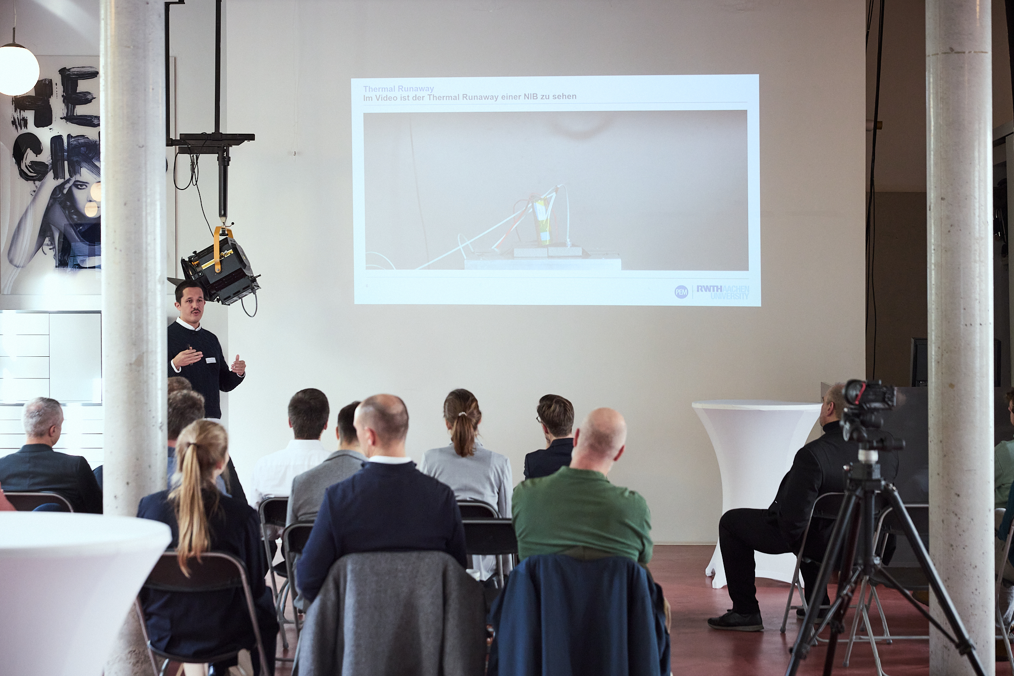 Ein Sprecher präsentiert vor einer Gruppe von Zuhörern, die auf Stühlen sitzen, und zeigt eine Projektion über 'Thermal Runaway' an der Wand. Im Hintergrund ist ein Kamera-Setup zu sehen.