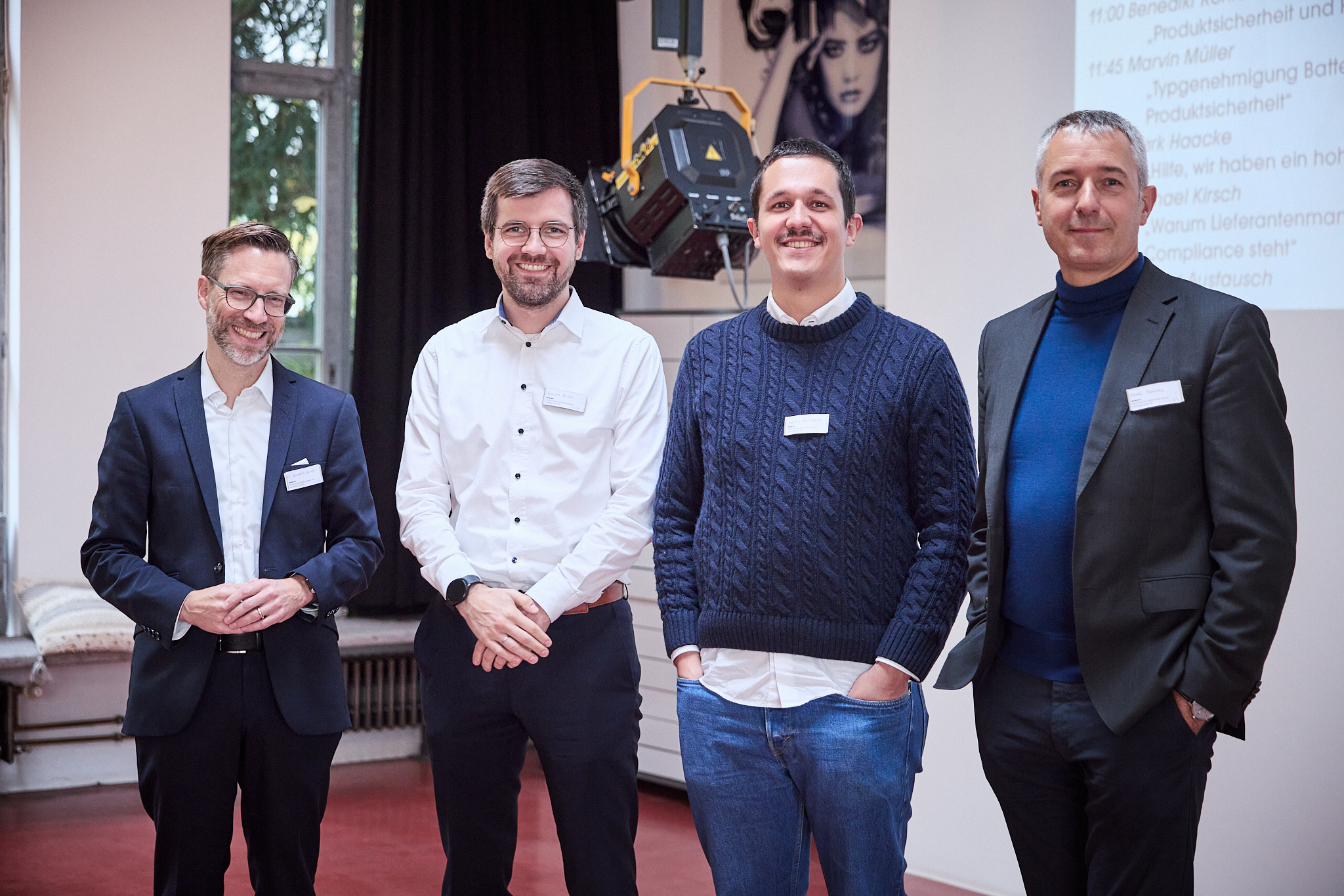 Vier Männer stehen in einem Raum. Der Mann links trägt einen Anzug und eine Brille. Neben ihm steht ein Mann im weißen Hemd mit Brille. Der dritte Mann trägt einen dunkelblauen Pullover und Jeans. Der Mann rechts hat einen dunklen Anzug an. Im Hintergrund ist ein Fenster und ein Bildschirm mit teilweise lesbarem Text.